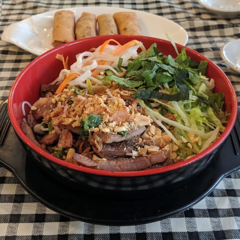 A red bowl of Vietnamese bun with meat, vegetables, and peanuts, with spring rolls in the background.